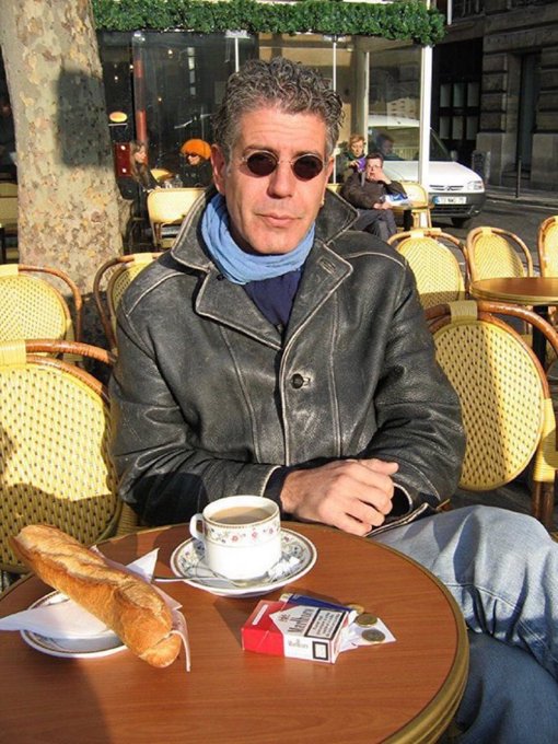 A man in sunglasses and a leather jacket sitting at an outdoor cafe table with coffee, a baguette, a sugar packet, and a cigarette pack.