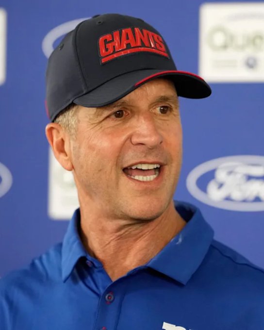 The caption should reflect a person that aligns with the post's subject. Let me provide a general caption without identifying the specific person.

A man wearing a black "GIANTS" cap and blue polo shirt smiles while speaking at what appears to be a Ford press conference, set against a backdrop with sponsor logos.