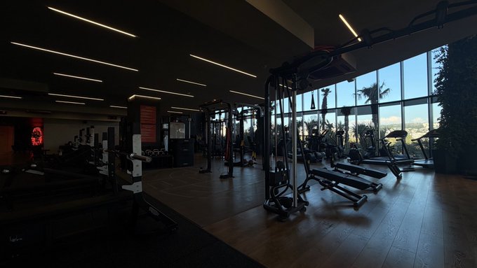 A dimly lit, spacious gym interior features weight machines and treadmills silhouetted against bright natural light coming through large windows.