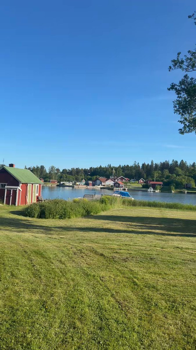 The image shows a beautiful view of a lake surrounded by grassy fields, with several boats floating on the water's surface. In the background, there are houses visible along the shoreline. The sky above appears blue, suggesting that it might be a clear day or during the summer season when people tend to spend more time outdoors enjoying various activities such as boating and picnicking by the lake.
