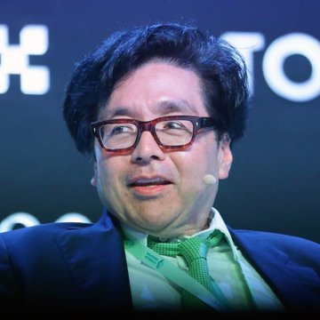 A close-up portrait of a man with dark hair and glasses wearing a dark suit, light shirt, and green tie.