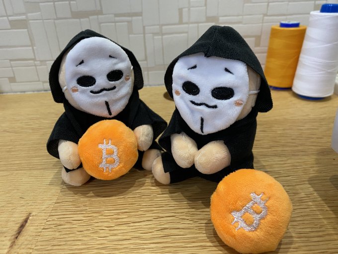 Two plush dolls wearing black hooded robes and white Anonymous/V for Vendetta-style masks sit on a wooden surface, accompanied by two orange plush coins featuring the Bitcoin "B" logo.