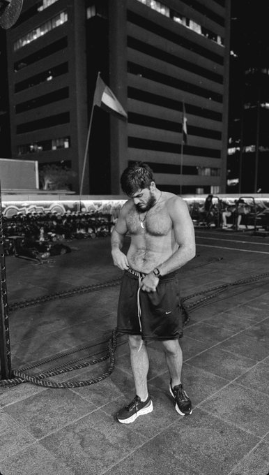 A black-and-white photograph of a shirtless, bearded man in athletic shorts and sneakers preparing for a battle ropes workout on an outdoor plaza at night, with tall city buildings in the background.