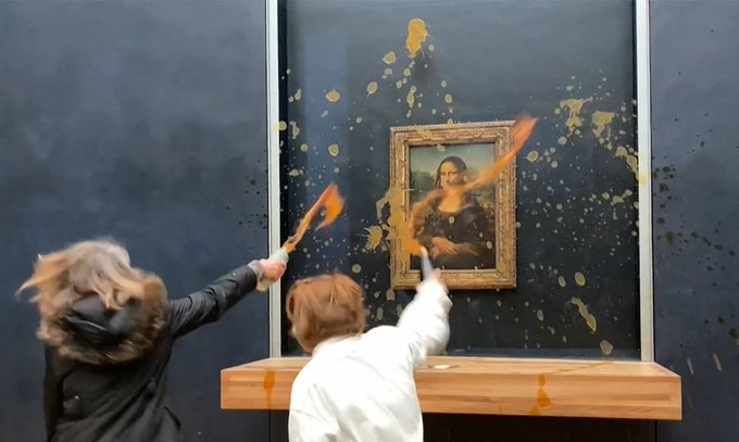 Two women spray orange liquid from spray bottles at the Mona Lisa painting in what appears to be an art protest, covering the artwork in splatters.