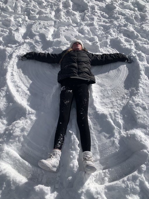 A person lies in the snow with arms outstretched, having made a snow angel impression around their body. They are wearing a black jacket, black pants, white boots, and a white beanie.