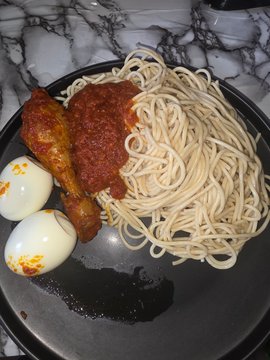 A plate of spaghetti with tomato sauce, fried chicken drumsticks, two boiled eggs, and what appears to be a piece of dark seaweed or garnish.