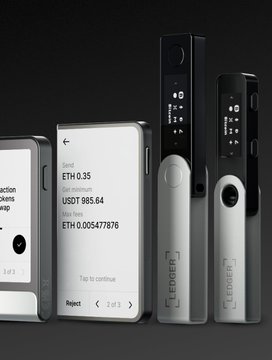 Three Ledger hardware wallets stand next to a smartphone displaying an Ethereum transaction of 0.005477876 ETH worth USDT 985.64.