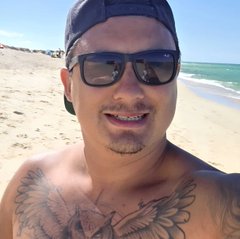 A shirtless man wearing a black cap and sunglasses smiles for a selfie on a sunny beach, with a chest tattoo visible.