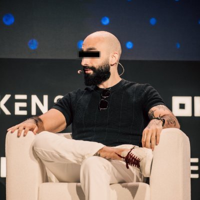 The image shows a bald man wearing sunglasses sitting in an armchair with his legs crossed. He appears to be seated at a podium or stage, possibly giving a speech or presentation. The background features a black wall with white dots, which could be part of a stage setup for an event. There is also text visible on the wall that reads "For some reason, I don' expect Mert to do something like that, I mean, to tweet that he sold all his zcash:native." This suggests that the man might have recently tweeted about selling all his zcash coins or a similar topic of interest.