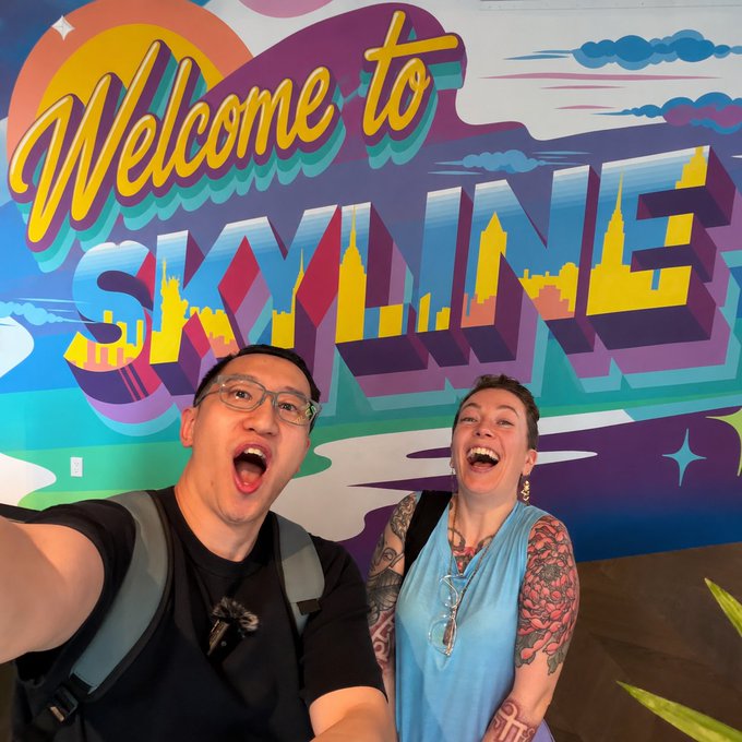 A selfie of a man and woman smiling excitedly in front of a colorful mural that reads 'Welcome to SKYLINE' in retro-style lettering.
