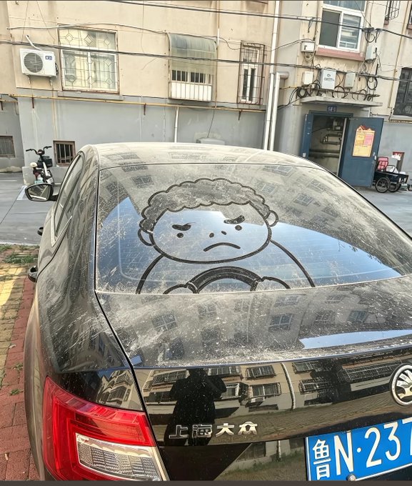 A black Volkswagen car parked outside an apartment building features a hand-drawn cartoon character on its dusty rear window depicting an angry-looking figure, with a Chinese license plate partially visible reading '鲁N·23'.