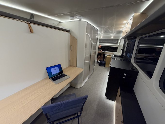 The interior of a customized van or camper featuring a long wooden desk with an open laptop on the left, a blue folding chair, and a kitchen area on the right.