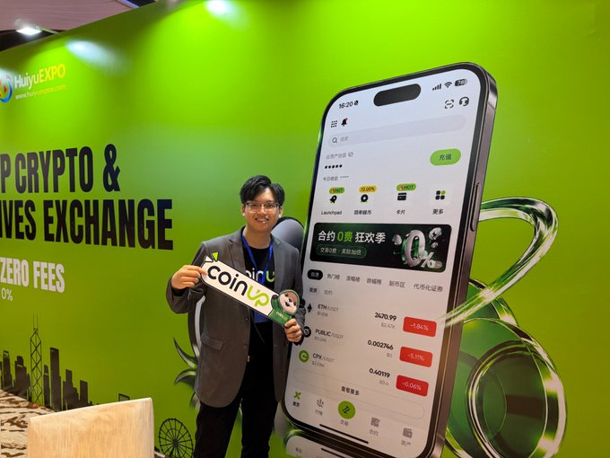 A man wearing glasses and a dark jacket holds a 'coinup' sign in front of a bright green 'HuiyEXPO' promotional backdrop featuring a giant smartphone displaying a crypto exchange app (ETH, TURBO, CSX).