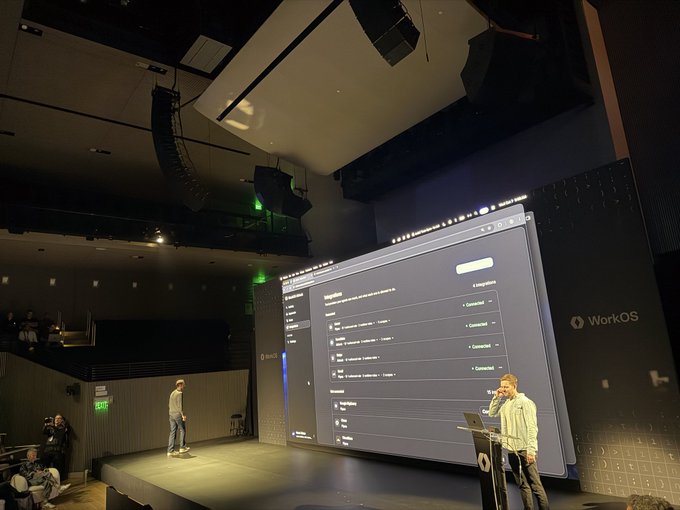 Two men present on a stage in a dim auditorium with a large screen displaying a WorkOS admin interface showing user management fields and connection settings.