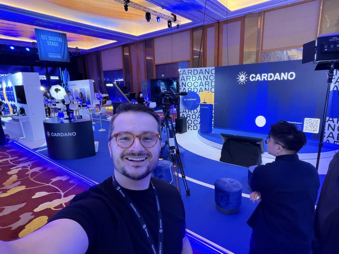 A man wearing glasses and a black CARDANO-branded lanyard takes a selfie at an indoor conference featuring prominent 'CARDANO' signage on the stage and 'BIT DIGITAL STAGE' displayed on a screen.