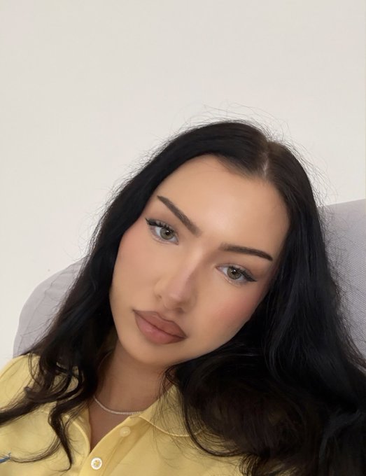 A selfie of a young woman with long dark wavy hair, wearing a yellow shirt, a delicate necklace, and makeup.