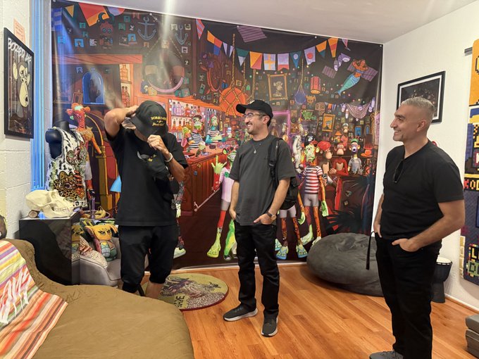 Three men stand in a colorful, art-filled room; one on the left wears a black cap and black shirt, adjusting his hat, while the other two, also in dark clothing, smile beside him.