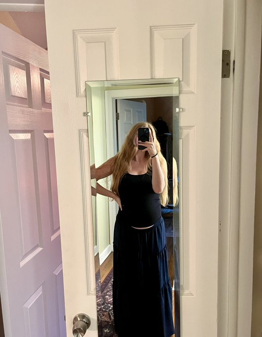A pregnant woman takes a mirror selfie in a black tank top and dark flowing maxi skirt, proudly showcasing her baby bump in what appears to be a bedroom. The image perfectly captures the celebratory "happy bump day" sentiment, highlighting the glow of pregnancy.