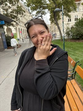 A woman stands outdoors in a black hooded jacket over a black dress, smiling and covering her mouth with her hand while displaying a ring, with buildings and a park bench visible in the background.