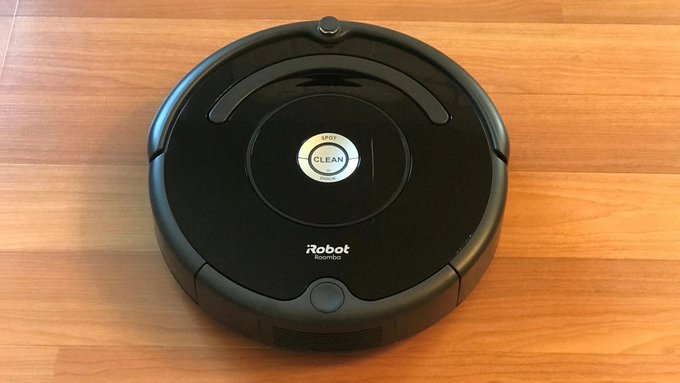 A black iRobot Roomba robotic vacuum cleaner sits on a wood floor, its central button illuminated and displaying the word 'CLEAN'.
