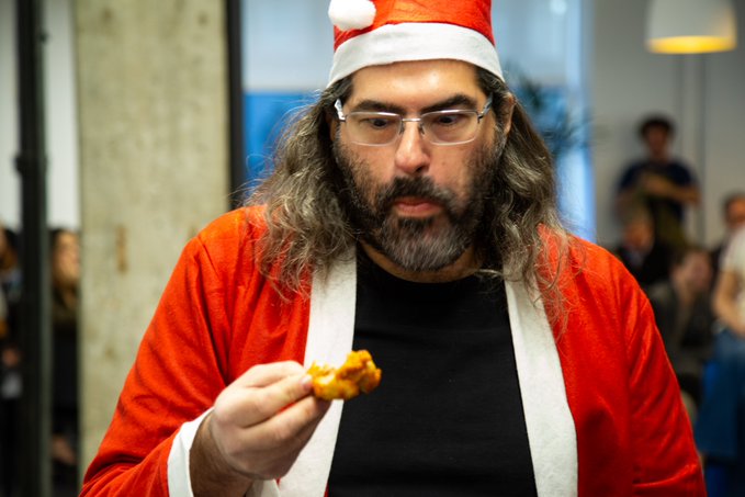 A man dressed in a Santa Claus costume, complete with a red hat, red jacket with white trim, and a long gray beard, is holding and examining a small piece of food while looking down at it. The festive outfit paired with the casual setting and the focus on the food suggests a holiday-themed gathering or party, aligning with the post's playful "blessings from GOD" theme featuring David Schwartz, Ripple's CTO.
