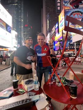 Two men stand side-by-side in a busy, illuminated city square at night, with one wearing a red and blue jersey displaying the text "banter" and the other in a black shirt, posing behind a red table surrounded by bright advertising billboards including one that reads "UNLEASH YOUR POWER".