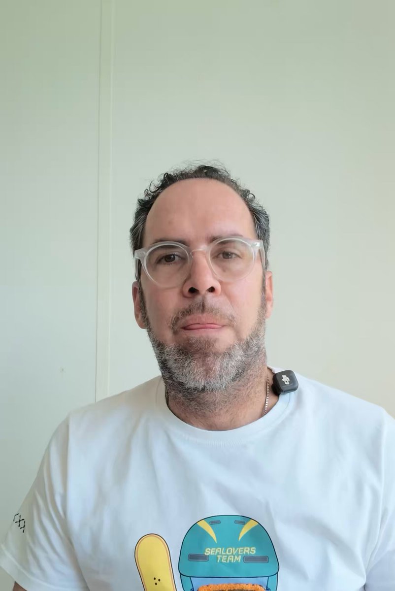 The image features a man wearing glasses and a white t-shirt with a cartoon design of a snowboarder. The man appears to be posing for a photo or selfie, possibly on social media platforms like Instagram. The caption provided about real-world assets moving from narrative to infrastructure is unrelated to the main subject matter of the image and does not provide any relevant information about the post.