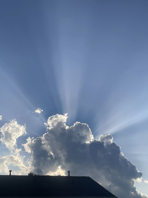 A dramatic photograph of sunbeams radiating outward from behind a large cumulus cloud above the dark silhouette of a building rooftop.