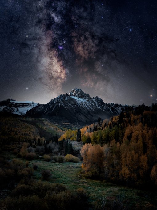 A breathtaking nighttime landscape featuring the Milky Way galaxy illuminating the sky above a snow-capped mountain peak, with a valley of autumn-colored trees in the foreground.