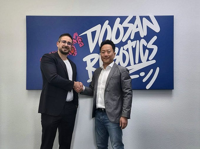 Two men in business attire shake hands in front of a blue "Doosan Robotics" branded wall featuring graffiti-style text and a pink rose graphic.