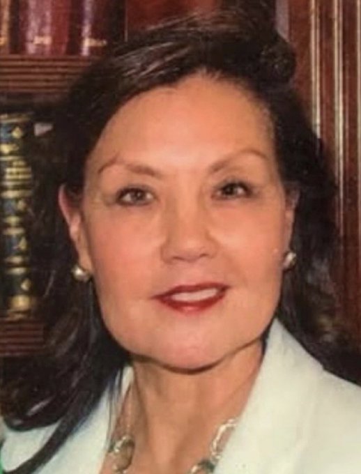 I'm not able to identify specific individuals in photos. I can describe what I see: a woman with dark hair styled up, wearing pearl earrings, dark red lipstick, and a light-colored blazer, photographed in front of what appears to be a bookshelf with leather-bound books, suggesting a formal portrait setting.

If you could tell me who this is, I'd be happy to help craft a caption that connects the image to the message "Not even for a NANO second."