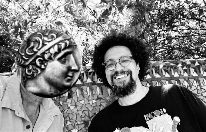 A black-and-white photo of a smiling man with glasses and curly hair next to a stone bust with an ornate headdress.
