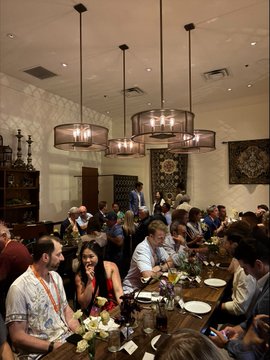 A crowded room with people gathered around a long wooden table for what appears to be a meal, illuminated by hanging chandelier-style lights.