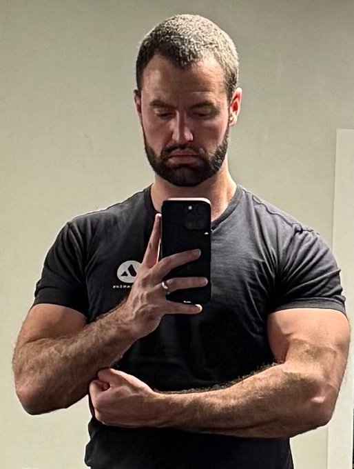 A muscular, bearded man wearing a black t-shirt takes a mirror selfie with a smartphone, looking down at the screen.
