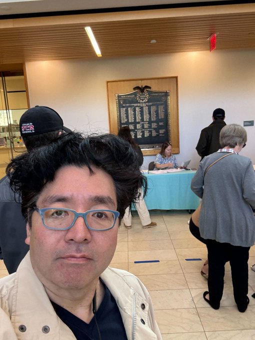 The image shows a man wearing glasses with his face partially visible, standing in front of a sign that says "Lately, Tomri'S hair has started to get messy again. $eth bmnr". The photo captures him posing for the camera while he is surrounded by other people who are also present in the scene.