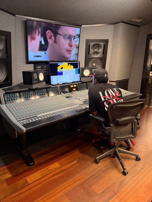 An audio engineer in a "LOVE" graphic t-shirt works at a large mixing console in a professional recording studio, with studio monitors and a TV displaying a man's face visible in the background. The image ties to the "Agent in the studio" caption by showing the behind-the-scenes work happening before the upcoming "AMP cypher" release.