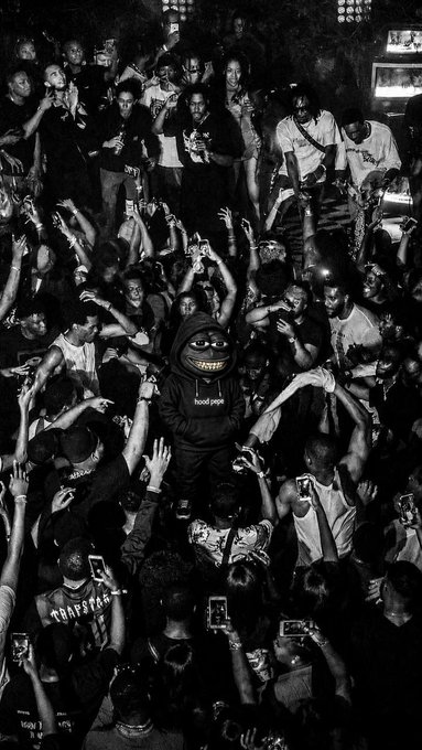 A black and white photograph of a person in a hooded sweatshirt with a grinning mask in the center of a crowd, with many hands raised and people holding up phones.