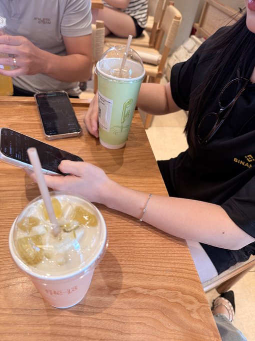 The image shows a close-up of two women at a wooden café table with iced matcha drinks, a phone, and a watch, while the context describes a conversation where a VIP client asks an "Alice" why she should switch from exchange B—prompting a targeted response highlighting lower fees, cashback rewards, and personalized service.