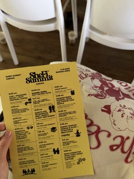 A hand holds a yellow "SheFi Summit Season 17 Scholar" brochure over a tablecloth featuring the Marilyn Monroe "Happy Birthday Mr. President" design, visually celebrating the user's acceptance into the SheFi scholars program and their journey from attending the summit to joining its first cohort.