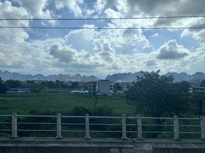 A scenic landscape view from behind a bridge railing, showing green fields, buildings, distant karst mountains, and a bright sky with scattered clouds.