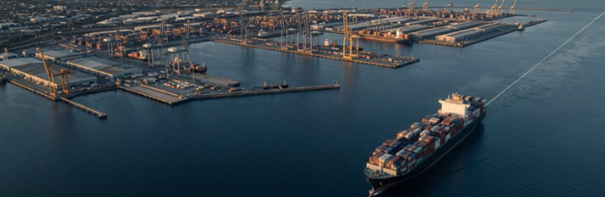 Aerial photograph of a large container ship sailing out of a commercial shipping port at sunset, with docks, cranes, and stacked shipping containers visible.