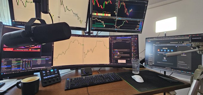 A trader's workstation setup with multiple monitors displaying stock/crypto charts, a microphone, keyboard, mouse, and a coffee mug on the desk.