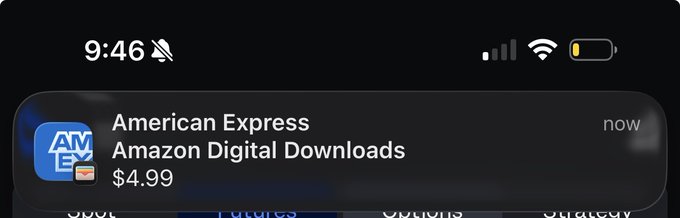 A smartphone notification from American Express for an Amazon Digital Downloads charge of $4.99 is displayed on a dark screen. The notification humorously contrasts with the post about being "homeless," showing how financial alerts still arrive even when juggling multiple temporary living situations.