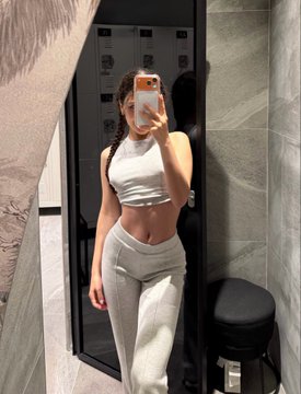 A mirror selfie shows a woman wearing a white halter crop top and white ribbed pants, holding a phone in a bathroom setting.