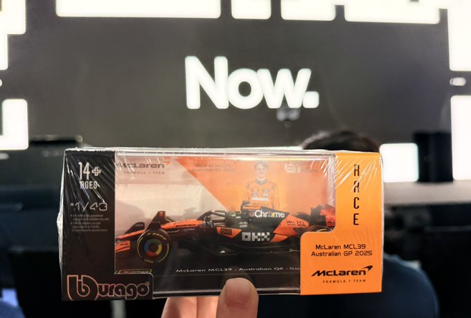 A person holds up a packaged Bburago 1:43 scale model of the McLaren MCL39 Formula 1 car used in the Australian GP 2025, with a 'Now.' sign in the background.