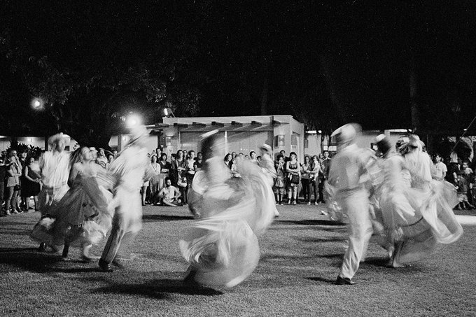 A black-and-white motion-blurred photograph shows dancers in flowing white garments performing outdoors at night while a crowd watches.