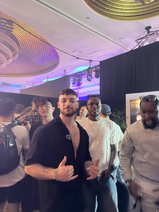 A casually dressed group poses at a crowded indoor event under purple lighting, with one man in the foreground making a hand gesture. The post’s “GM” message suggests a good-morning greeting, fitting the photo’s upbeat social setting.