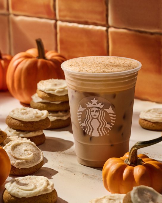A Starbucks iced coffee drink is surrounded by pumpkin cookies, mini pumpkins, and cinnamon sticks against a tiled wall background.