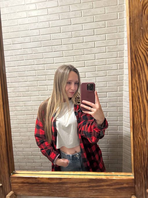 A young woman with long blonde hair takes a mirror selfie wearing a red and black flannel shirt over a white t-shirt and jeans.