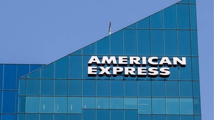 A modern glass skyscraper displays the "AMERICAN EXPRESS" logo prominently against a clear blue sky, visually illustrating the breaking news of American Express's reported global payments partnership with Ripple involving XRP.